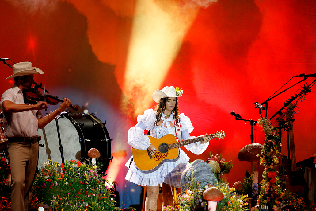 music artist sierra ferrel performing with guitar on wvu event stage surrounded by mushrooms and greenery